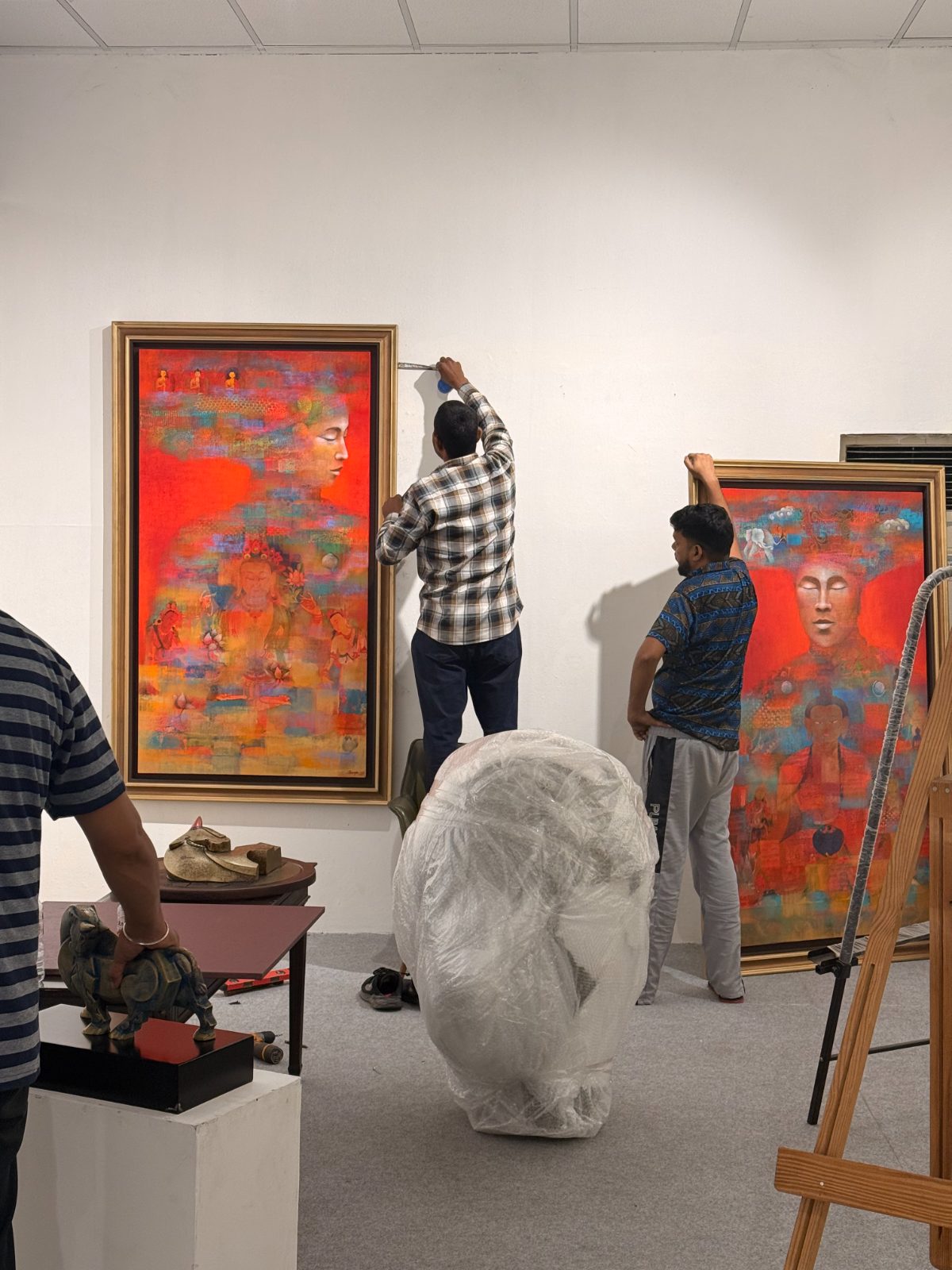 Two installers hanging tall red canvases, a sculpture wrapped in plastic nearby